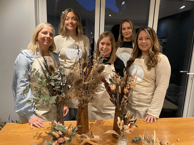 Five of the Bouquet Cards team in Dried Blooms aprons behind a workbench of dried flower arrangements